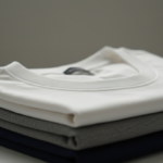 A neat stack of three folded t-shirts with a soft white crew neck shirt on top, resting above grey and dark navy blue layers.